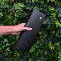 Image of a female hands holding a rolled-up earthing mat next to a wall of lush plants. The vibrant plant backdrop highlights the theme of natural grounding, presenting the mat as a practical accessory for connecting with nature