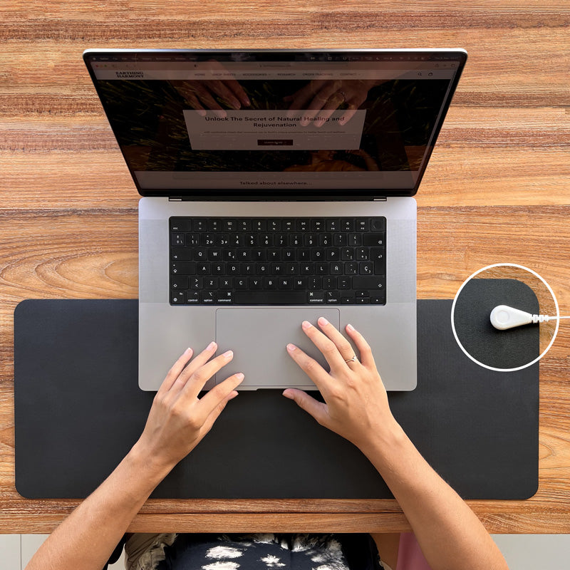 Woman working on macbook computer protecting herself from EMF with Earthing mat that is placed on natural wooden desk